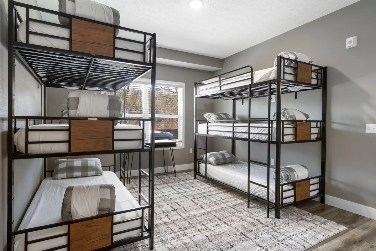 A cozy bunk room featuring multiple built-in bunk beds, a window letting in natural light, and a soft rug on the floor. The space is designed for group fun and late-night chats, creating a welcoming adventure zone for kids or groups.