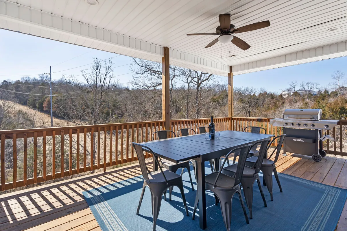 An outdoor dining area on the upper deck, complete with a dining table, grill, bottle of wine, and surrounded by trees. The space invites guests to enjoy sunset dinners, barbecues, or peaceful breakfasts in the fresh mountain air.