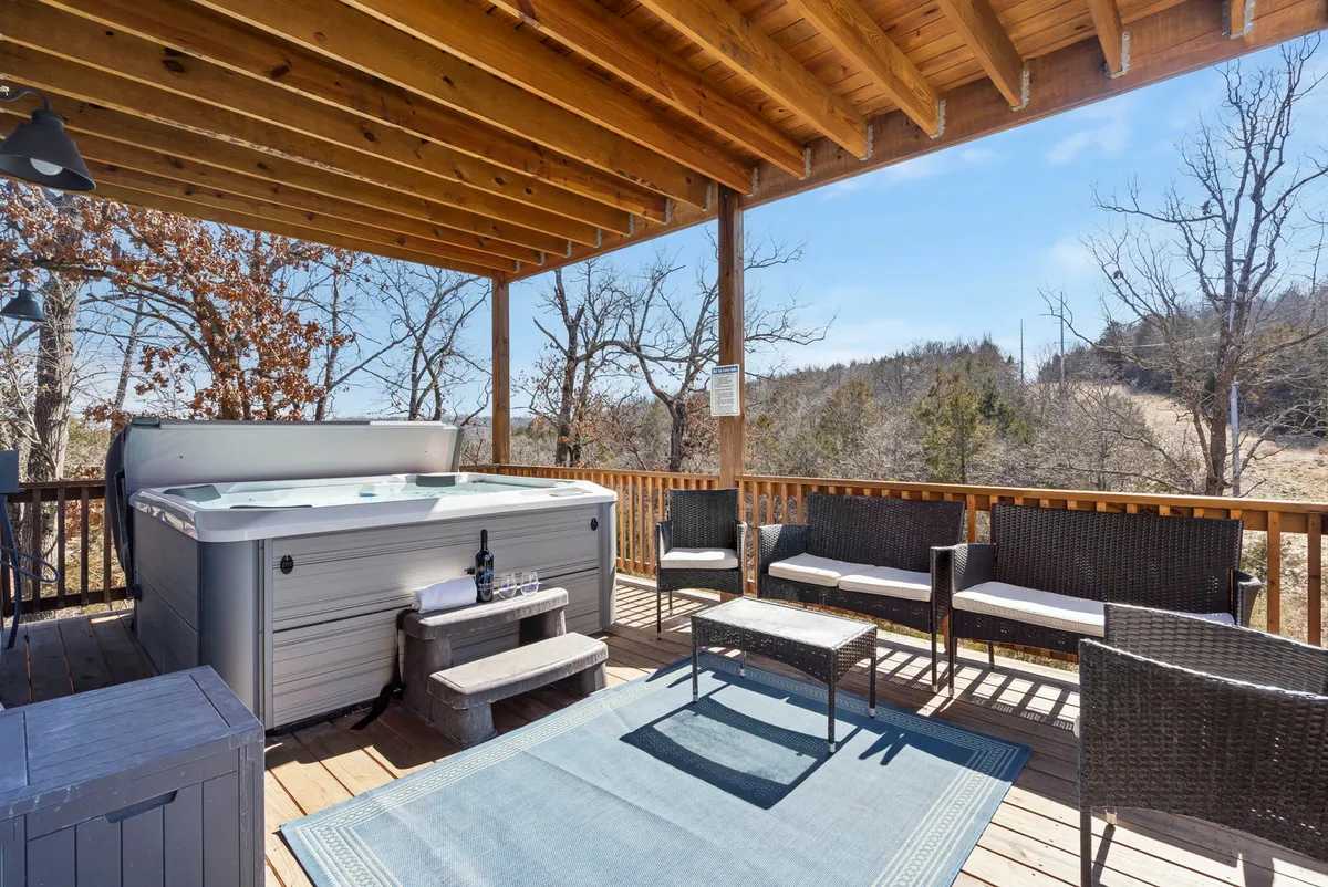An inviting exterior view featuring a wooden deck nestled among the trees, with a private hot tub and comfortable seating area overlooking lush natural landscape. The scene is set for relaxing and socializing outdoors, with space to take in the forested Ozark surroundings both day and night.