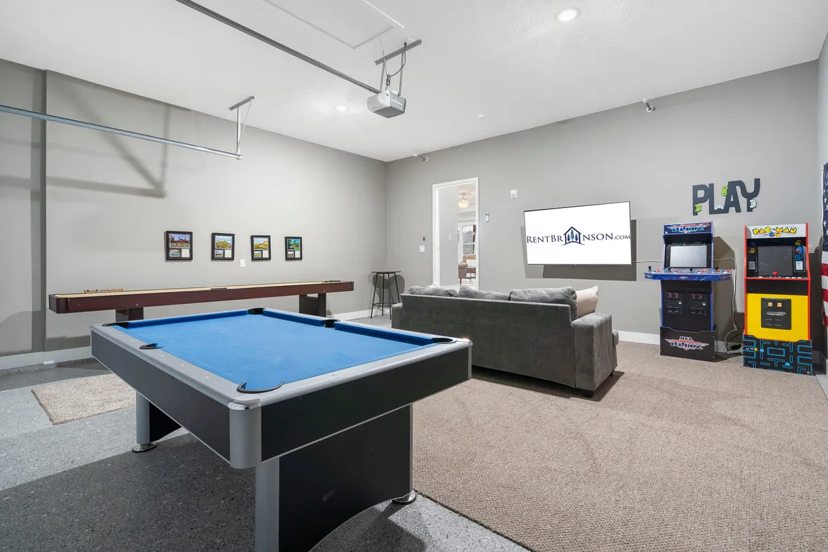 A spacious game room featuring a pool table, comfortable sofa, arcade games, and a large projector screen. Brightly lit and arranged for social gatherings, the space invites playful competition and group entertainment.