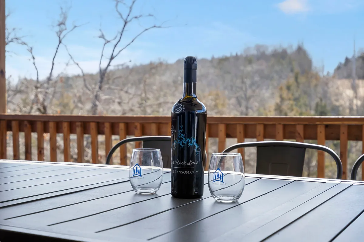 A close-up of a bottle of wine and two glasses on a wooden deck railing, with a sweeping view of the distant landscape at sunset. The mood is intimate and welcoming, suggesting an ideal spot for relaxing, sharing drinks, and conversation after a day of fun inside.
