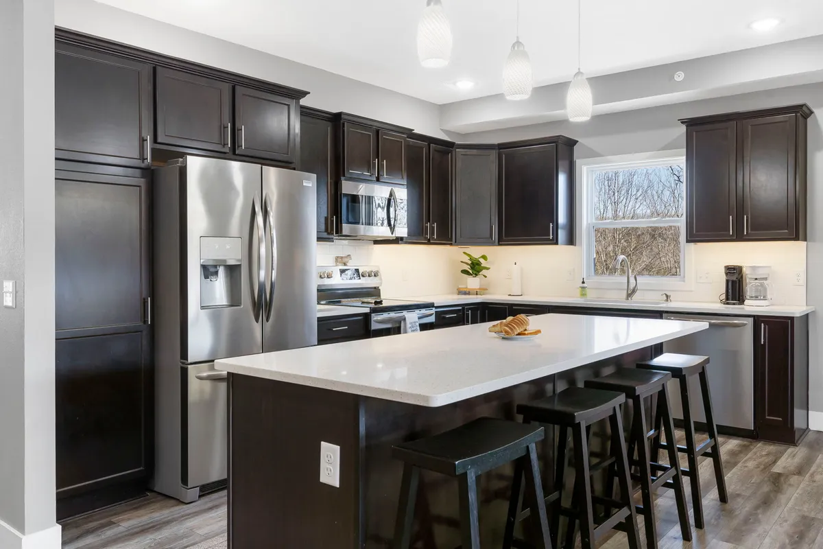 The image showcases a bright, modern kitchen with a spacious island countertop, rich dark wood cabinetry, gleaming stainless steel appliances, and elegant pendant lighting. This inviting space is perfect for gathering, sharing meals, and creating memories, all set within a stylish and comfortable environment.
