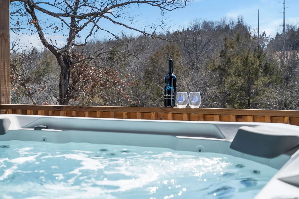The image showcases a tranquil outdoor hot tub setting, featuring a bottle of wine and two glasses staged on the tub’s edge. The natural surroundings and soft lighting evoke a sense of peaceful relaxation and a touch of luxury, ideal for unwinding after a day of adventure.