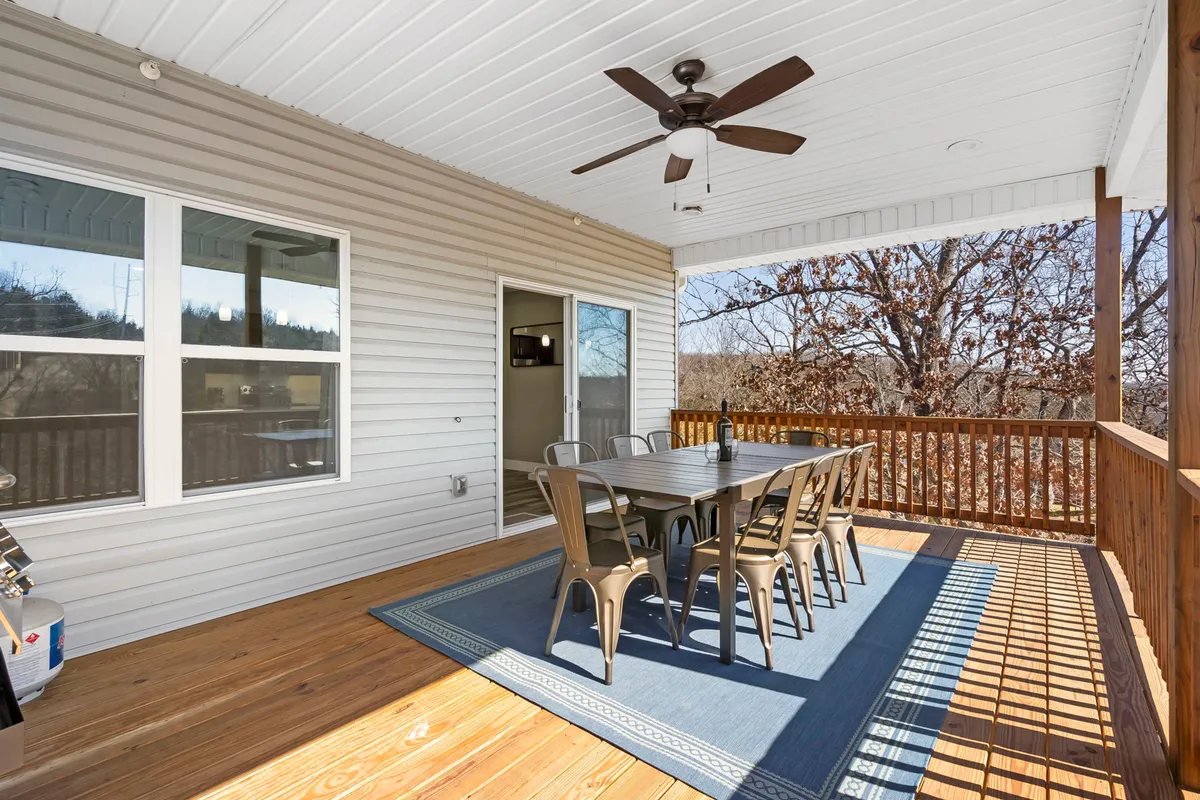 A spacious wooden deck set for outdoor dining, featuring a dining table, ceiling fan, large windows, and lush trees surrounding the area. The space showcases the opportunity for meals above the treetops, blending seamlessly with the woodland setting.