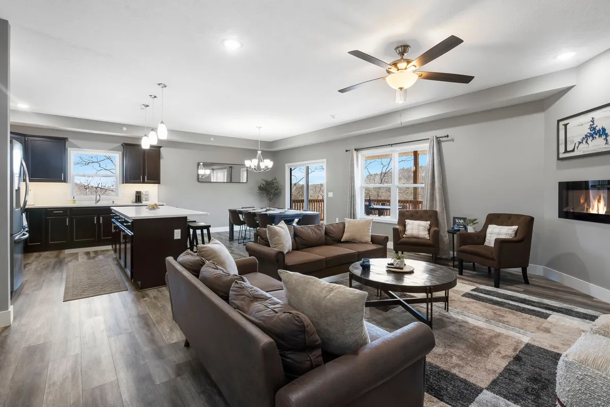 A welcoming, sunlit interior featuring a plush sofa, a spacious dining area, a kitchen island, and a modern fireplace. Large windows flood the open-plan space with natural light, creating an inviting atmosphere perfect for gatherings and cozy evenings.